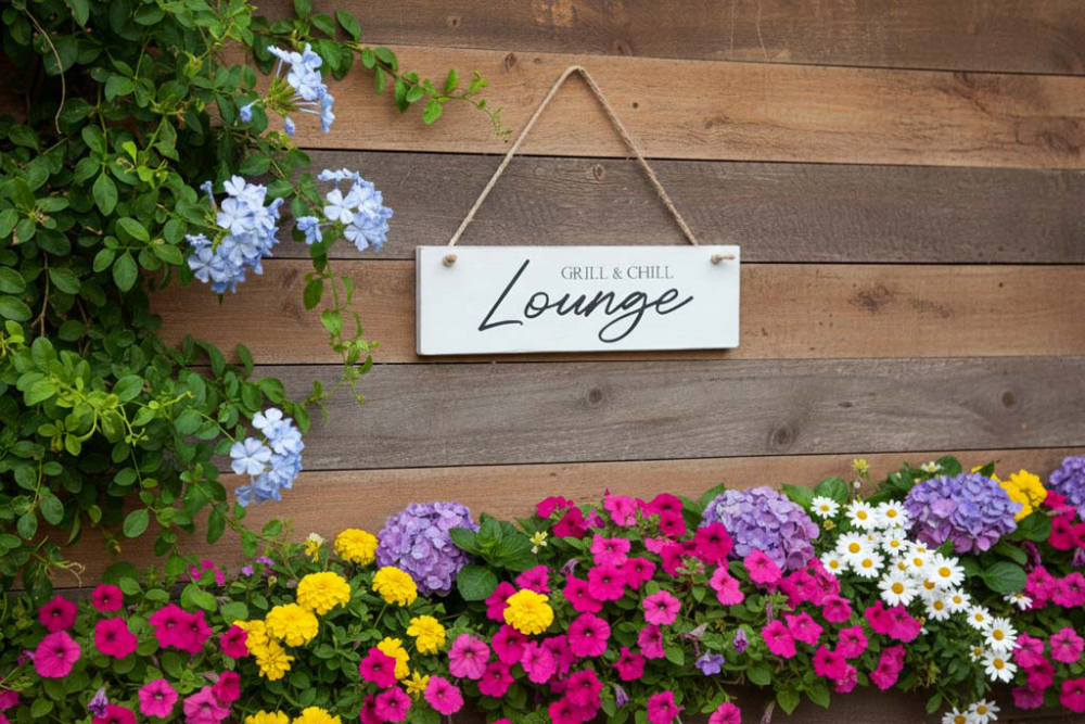 Deko-Schild Grill & Chill Lounge aus Holz, braun mit weißer Schrift, aufgehängt an einer rustikalen Holzwand im Gartenbereich als gemütliche Grillplatz-Dekoration